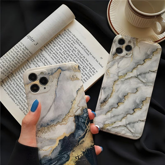 Two marble-patterned phone cases held by a hand with an open book and cup in the background.